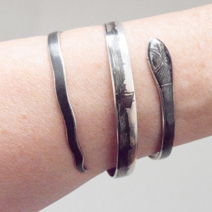 May include: Three silver bracelets on a wrist. One is a simple band, another has a landscape design, and the third is a snake head cuff. The bracelets are made of a shiny metal.