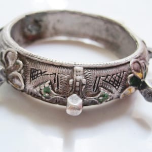 Peut inclure: Un bracelet argenté orné de motifs floraux et géométriques complexes. Le bracelet présente quatre ornements rectangulaires en relief et de petites zones de peinture verte et jaune. Le design suggère un style vintage, d'environ 6 cm de diamètre.