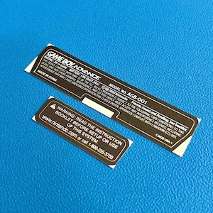 Może przedstawiać: Dwie prostokątne naklejki z tekstem, jedna długa i jedna krótka, na niebieskim tle. Dłuższa naklejka zawiera napis "GAME BOY ADVANCE" i dane techniczne. Krótsza naklejka ostrzega przed przeczytaniem instrukcji.