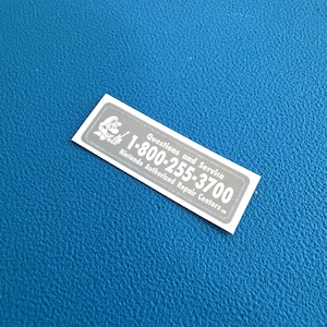 Może przedstawiać: Prostokątna srebrna naklejka z białą grafiką Mario, napisem "Questions and Service", numerem telefonu "1-800-255-3700" oraz tekstem "Nintendo Authorized Repair Centers" z małym symbolem znaku usługowego na końcu.
