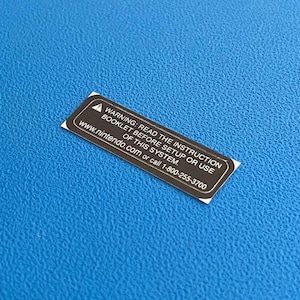 Op de afbeelding: Een rechthoekige zwart-witte sticker met de tekst "WARNING: READ THE INSTRUCTION BOOKLET BEFORE SETUP OR USE OF THIS SYSTEM. www.nintendo.com or call 1-800-255-3700". De sticker ligt op een blauw, getextureerd oppervlak.