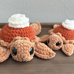 May include: Two handmade crochet turtle plushies with pumpkin pie tops. The turtles are light brown with large black eyes. The pumpkin pie tops are orange with white whipped cream. Autumn leaves are in the background.