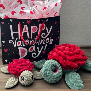 May include: Two handmade crochet turtle plushies with red rose shells and green flippers. A black gift bag with red handles and the text "Happy Valentine's Day" is in the background. The bag is decorated with hearts.