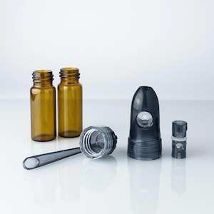 Puede incluir: Un surtido de botellas y componentes de vidrio de laboratorio o farmacia. Dos botellas de vidrio de color ámbar se encuentran junto a un componente cilíndrico transparente con un mango largo y delgado. También hay un componente redondeado de color gris oscuro y un componente pequeño y cuadrado.