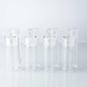 May include: A collection of small, clear glass vials with screw-on lids. The vials are cylindrical and stand upright, with a clear, minimalist aesthetic. The background is a plain white, emphasizing the vials' transparency and design.
