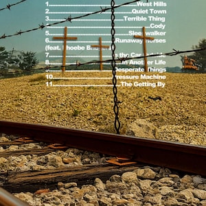 May include: Album cover for "Pressure Machine" by The Killers, featuring a landscape with barbed wire, wooden crosses, and railroad tracks. The tracklist includes "West Hills," "Quiet Town," and "Runaway Horses." The Island Records logo is in the upper left corner.