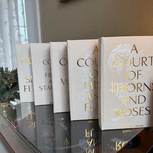 Pode incluir: Uma coleção de livros de capa dura com capas creme e letras douradas. Os títulos incluem "A Court of Thorns and Roses" e outros volumes relacionados. Os livros estão dispostos sobre uma superfície reflexiva.