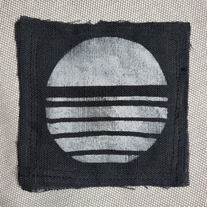 May include: A square, black fabric patch featuring a white, stylized sun design. The sun is depicted as a half-circle above horizontal lines. The patch has a stitched border and a textured appearance, suggesting a handmade item.