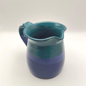 May include: A handmade ceramic pitcher with a handle. The pitcher is painted with a gradient of colors, transitioning from a deep blue at the base to teal and turquoise at the top. The handle is a darker shade of blue.