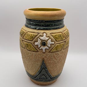 May include: A decorative ceramic vase with a textured, earthy brown surface. The vase features geometric patterns in yellow, black, and white, with a wide rim and a slightly flared base. The interior rim is a light green color.