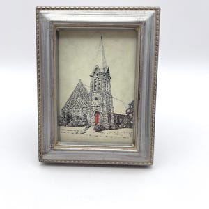 May include: A small, rectangular silver picture frame with a detailed black and white sketch of a church. The church has a tall steeple and a red door. The frame has a beaded edge.