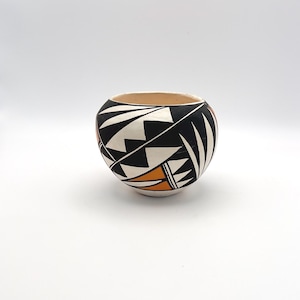 May include: A round, hand-painted ceramic bowl with a geometric design. The bowl features a black, white, and orange pattern on a cream-colored base. The interior is a light tan color.