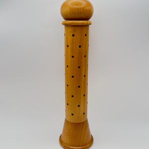 May include: A tall, wooden pepper mill with a light brown finish. The mill has a rounded top, a cylindrical body with evenly spaced holes, and a flared base. The pepper mill is standing upright against a white background.