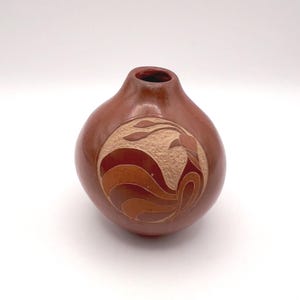 May include: A brown ceramic vase with a rounded body and a narrow neck. The vase features a carved design of a bird or stylized feather pattern in shades of brown and tan. The vase is approximately 6 inches tall.