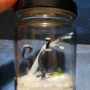 May include: A clear glass jar with a black lid, containing a miniature scene. Inside, there's a white sandy base, a white and blue structure, and several black, snake-like figures. A small green worm is also present.