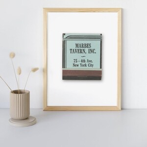 May include: Framed print of a vintage matchbook from Marbes Tavern, Inc., located at 75 - 4th Ave., New York City. The matchbook has a teal and brown colour scheme, and is displayed in a natural wood frame.