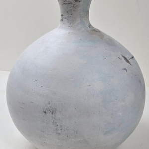 May include: A round, light gray vase with a narrow neck and a wide, bulbous body. The vase has a textured surface with visible imperfections and subtle blue and black accents, suggesting an aged appearance. The vase is likely made of ceramic or a similar material.