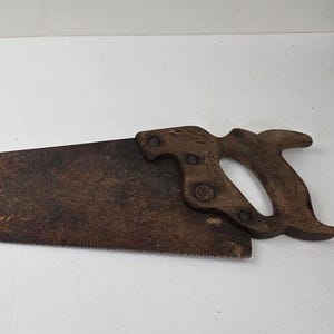 Puede incluir: Una sierra de mano antigua con un mango de madera y una hoja dentada y oxidada. La hoja de sierra mide aproximadamente 61 cm de largo. El mango es de color marrón oscuro y tiene un diseño curvo para el agarre. La sierra probablemente se utiliza para trabajar la madera.