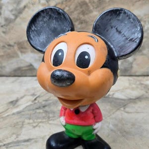 Puede incluir: Una figura vintage de Mickey Mouse. La figura tiene cabeza y orejas negras, cara naranja y camisa roja con pantalones verdes. La figura est&aacute; de pie sobre una superficie de color claro.