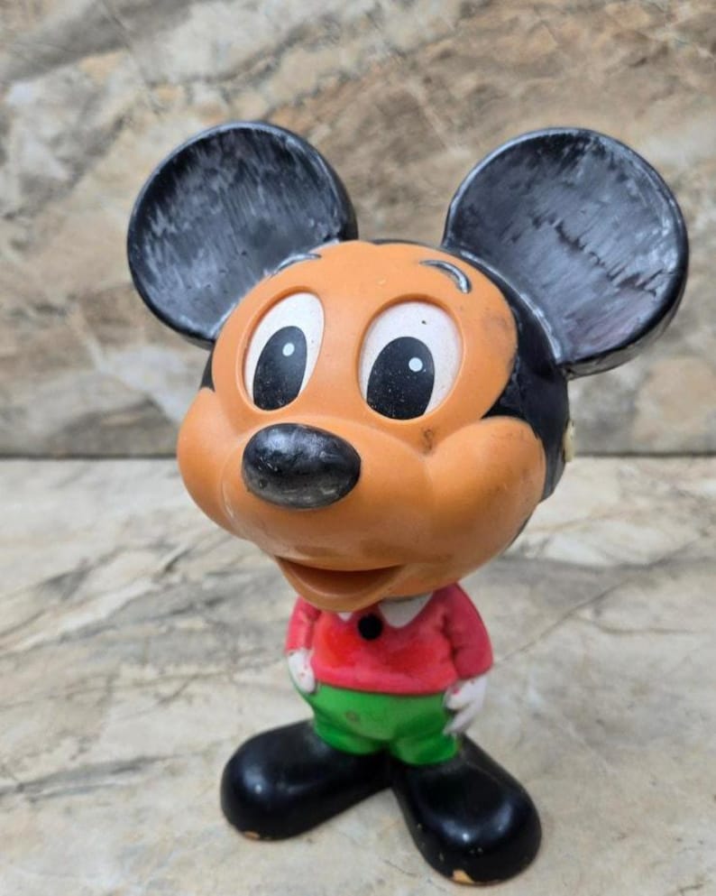 Puede incluir: Una figura vintage de Mickey Mouse. La figura tiene cabeza y orejas negras, cara naranja y camisa roja con pantalones verdes. La figura est&aacute; de pie sobre una superficie de color claro.