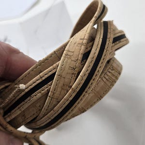 Puede incluir: Primer plano de una correa de corcho enrollada con una franja negra. La correa está hecha de material de corcho natural con grano y textura visibles. La franja negra recorre la longitud de la correa, agregando un elemento de diseño contrastante.