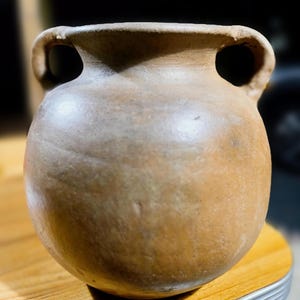 Puede incluir: Un jarrón de cerámica marrón hecho a mano con dos asas de bucle. El jarrón tiene un cuerpo redondeado y una abertura ancha. La superficie tiene un acabado mate y una apariencia natural y terrosa. El jarrón probablemente se utiliza con fines decorativos.
