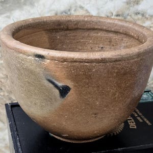 Peut inclure: Un bol rond en céramique marron fait à la main avec un large bord. Le bol a une surface légèrement texturée et un motif sombre et abstrait sur le côté. Le bol est posé sur une surface noire.