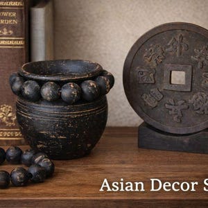 May include: An Asian decor set featuring a dark, weathered vase adorned with dark beads, a decorative coin, and a bracelet of beads. The set is displayed on a wooden surface with the text "Asian Decor Set".