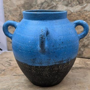 Könnte beinhalten: Eine blau-schwarze Keramikvase mit zwei Schlaufengriffen an den Seiten und einem an der Vorderseite. Die obere Hälfte der Vase ist in leuchtendem Blau bemalt, während die untere Hälfte schwarz ist. Die Vase hat ein rustikales, strukturiertes Aussehen.