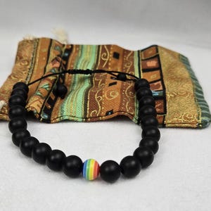 Puede incluir: Pulsera de cuentas negras con una sola cuenta de color arcoíris. La pulsera está sobre una bolsa de tela estampada con cierre de cordón. Las cuentas son mate y la cuenta arcoíris añade un toque de color.