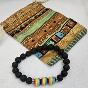 Puede incluir: Pulsera de piedra de lava negra con tres cuentas a rayas arcoíris. La pulsera se muestra con una bolsa con cordón estampada en tonos marrón, verde azulado y dorado. La bolsa tiene un cordón de color crema.