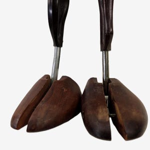 May include: A pair of dark brown wooden shoe trees featuring a split-toe design, a central metal rod, and a dark brown plastic handle at the top. The wooden components are shaped to fit inside footwear to maintain their original form and structure.