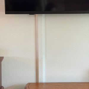 May include: A white wall with a textured surface. A white cable cover runs vertically down the wall. A dark television is mounted at the top of the wall. A wooden cabinet is at the bottom left.