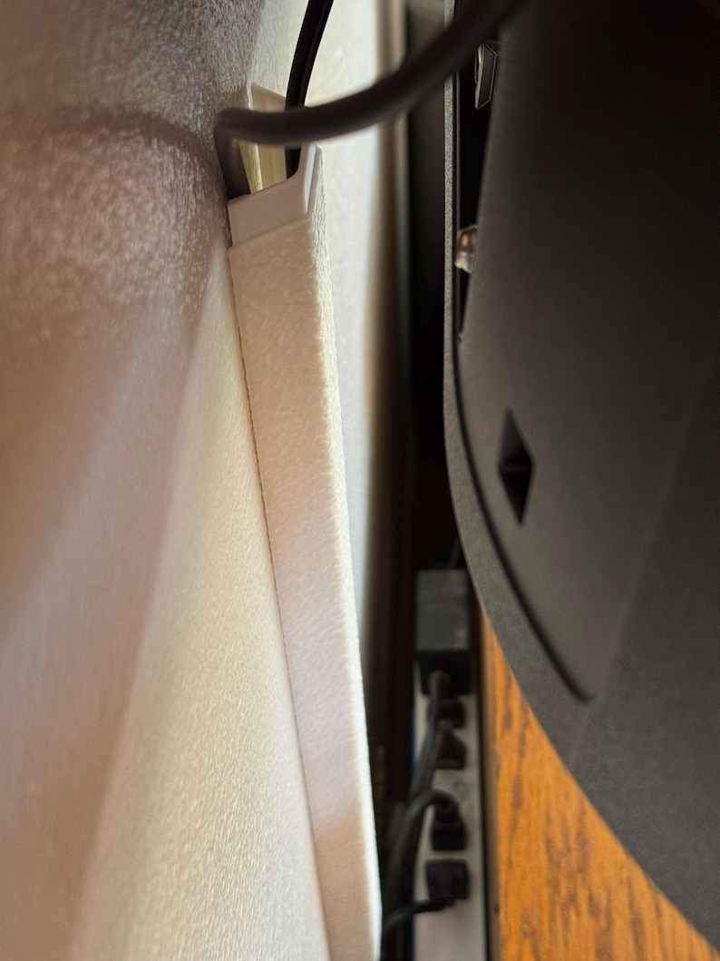 May include: A close-up view of a white cable management system installed on a wall. A black cable is routed through the system, alongside a black TV. The wall has a textured surface.