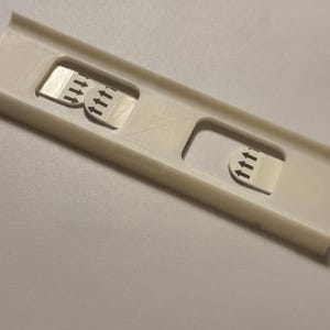 May include: A white plastic device with three rectangular cutouts. Each cutout contains a small, rectangular sticker with three upward-pointing black arrows. The device has a rectangular shape with raised edges.
