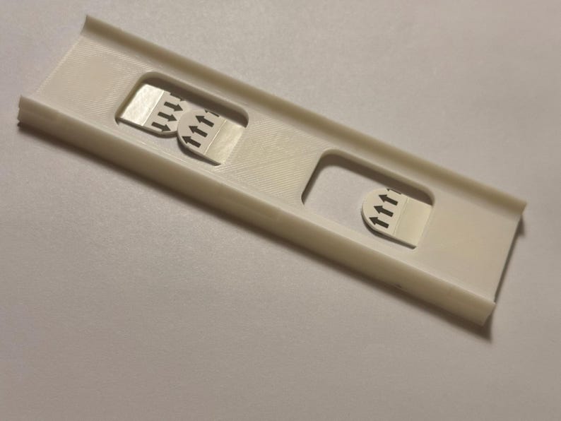 May include: A white plastic device with three rectangular cutouts. Each cutout contains a small, rectangular sticker with three upward-pointing black arrows. The device has a rectangular shape with raised edges.