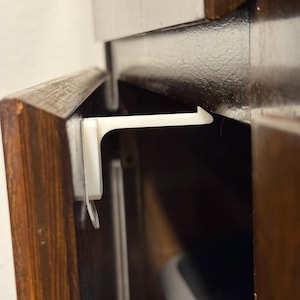 Puede incluir: Un cierre de seguridad de armario de plástico blanco instalado en un armario de madera marrón oscuro. El cierre está diseñado para evitar el acceso al contenido del armario. El armario tiene un acabado brillante.