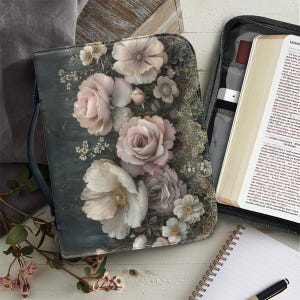May include: A floral Bible cover with a dark teal background and a design of pink and white roses. The cover is open, revealing a Bible and a pen. A notebook and pen are also visible.