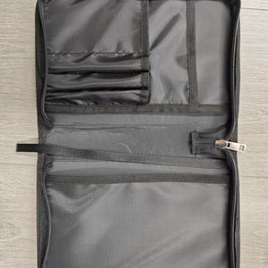 May include: Open, rectangular black travel organizer with multiple interior pockets and elastic straps. The organizer has a full-length zipper closure and a small fabric loop. The interior is a lighter shade of black.