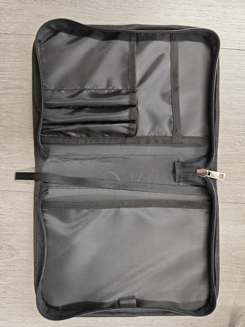 May include: Open, rectangular black travel organizer with multiple interior pockets and elastic straps. The organizer has a full-length zipper closure and a small fabric loop. The interior is a lighter shade of black.