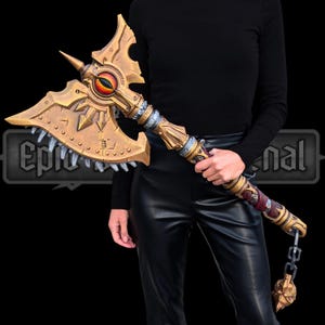 Puede incluir: Un hacha dorada grande y ornamentada con un diseño detallado, con un emblema central en forma de ojo con tonos rojos y naranjas. El hacha tiene una parte superior puntiaguda, un borde dentado y un mango decorativo con una cadena y un colgante de calavera. El texto "Epic nal" es visible.