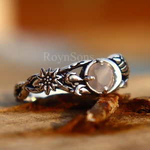 May include: A silver ring with a floral and sunburst design, featuring a light pink, round gemstone. The ring is set on a wooden surface, with the brand name "RoynSons" visible.