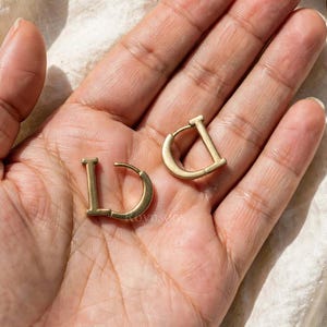 May include: A pair of gold-toned hoop earrings shaped like the capital letter D. Each earring features a straight vertical bar connected to a curved semi-circular base, creating a distinct geometric silhouette with a smooth, polished metallic finish.