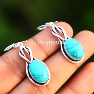 Puede incluir: Un par de pendientes plateados con piedras ovaladas de turquesa. Los pendientes tienen un diseño calado decorativo sobre la piedra. Las piedras de turquesa son de un color azul-verdoso brillante.