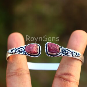 K&ouml;nnte beinhalten: Silberfarbenes Armreif-Armband mit zwei rohen, r&ouml;tlich-rosa Edelsteinen. Das offene Armband hat verzierte Details um die Steine. Die Edelsteine sind in eine dekorative Silberfassung gesetzt und schaffen ein einzigartiges Schmuckst&uuml;ck.