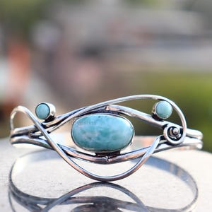May include: A silver cuff bracelet featuring a large, oval, light blue gemstone at the center, flanked by two smaller, round, light blue stones. The bracelet has a flowing, open design.