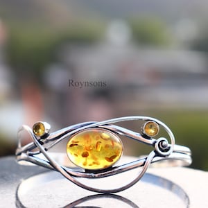 Peut inclure: Un bracelet en argent avec une pierre d'ambre ovale au centre et deux petites pierres jaunes de chaque côté. Le bracelet présente un design unique et fluide. Le mot "Roynsons" est visible en arrière-plan.