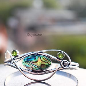 Può includere: Bracciale color argento con un elemento centrale ovale in conchiglia di abalone con iridescenze blu, verdi e gialle. Due gemme verdi accentuano il design. La parola "Roynsons" è visibile nell'immagine.
