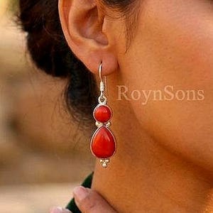 Puede incluir: Pendientes de plata con vibrantes piedras de coral rojo. Cada pendiente presenta un colgante de coral en forma de lágrima debajo de una cuenta de coral redonda más pequeña. El nombre de la marca "RoynSons" es visible.