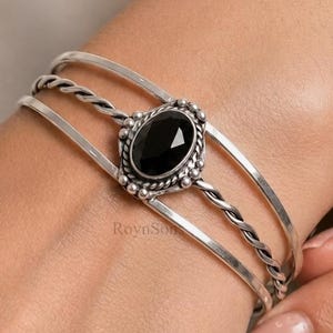 May include: A silver cuff bracelet featuring three distinct bands, including a central twisted rope design. The centrepiece is an oval-shaped, faceted black stone set in a decorative frame with small silver beads. The text RoynSon is visible.